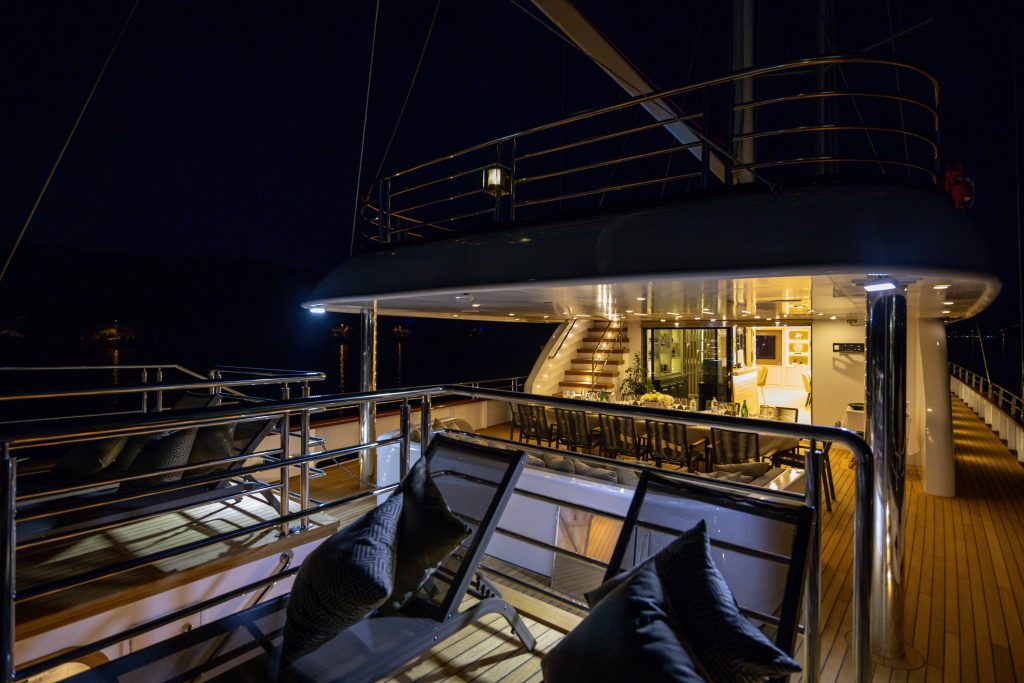 Luxurious yacht deck at night, featuring cushioned lounge chairs, polished railings, warm lighting, a dining area with a long table and chairs, and an illuminated staircase leading to the upper level.