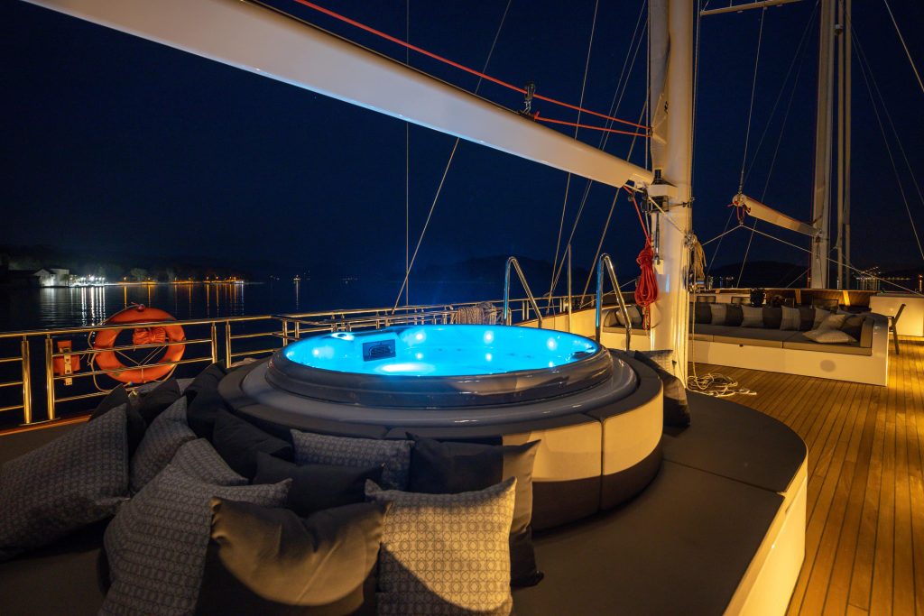 A luxury yacht deck at night features a glowing round hot tub surrounded by cushions. The deck is lit softly, with calm water and shoreline lights visible in the background.