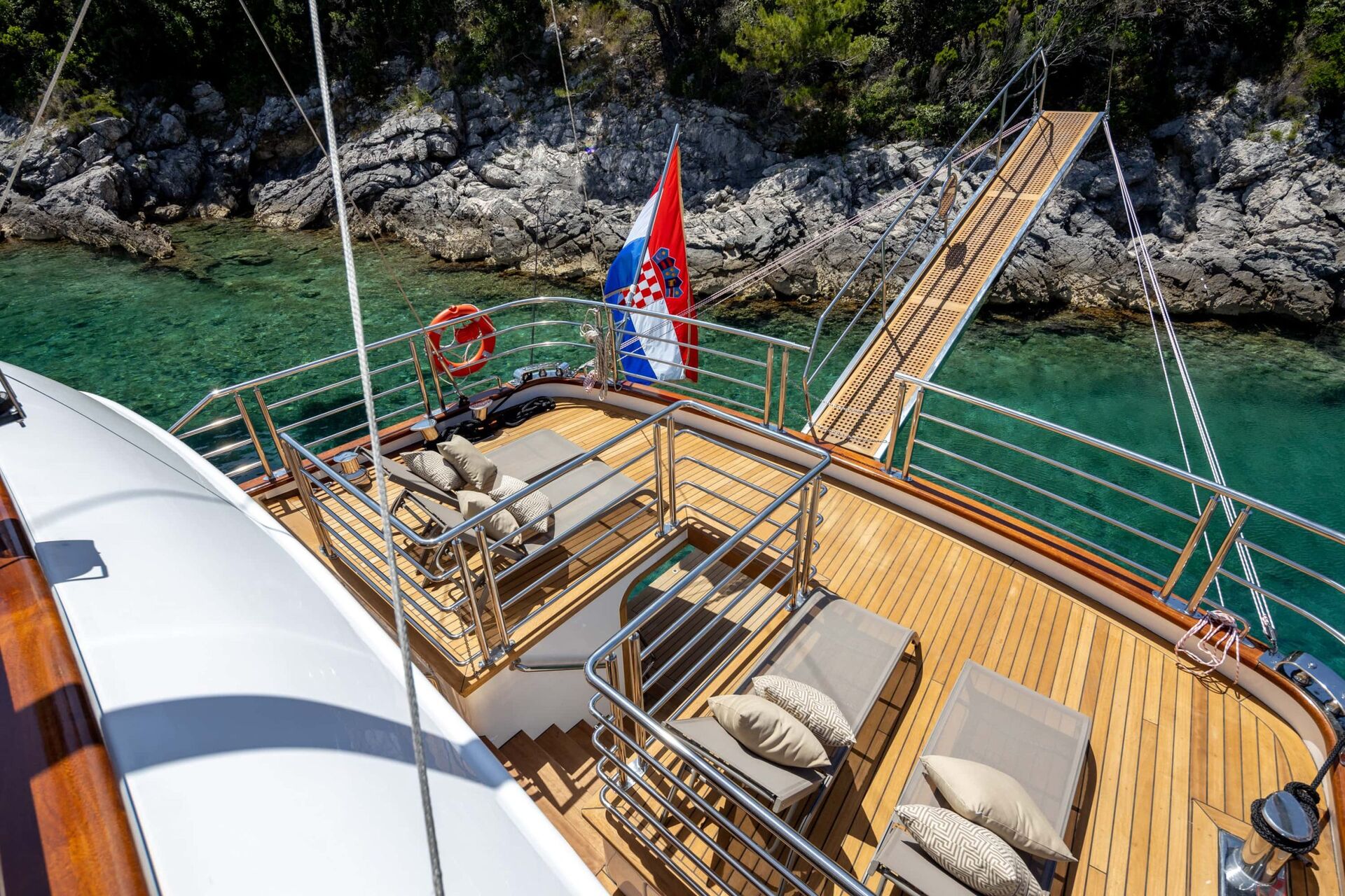 View from a yacht deck with sun loungers, a Croatian flag, a lifebuoy, and a gangway leading to rocky shore over clear turquoise water.
