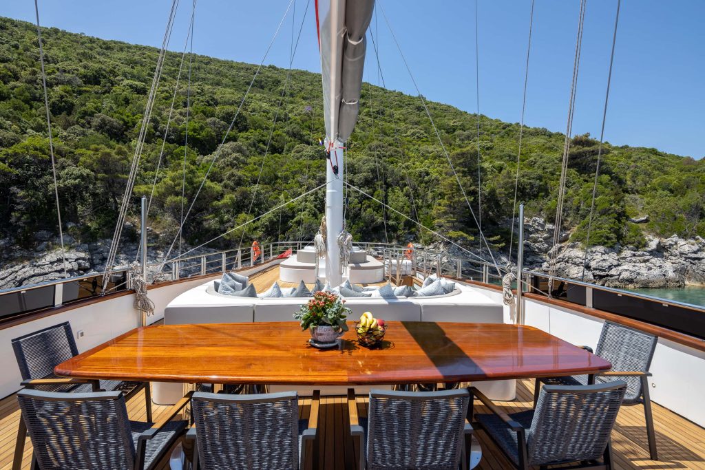 A polished wooden dining table with wicker chairs is set on the deck of a yacht, overlooking cushioned seating and surrounded by lush green hills and rocky shoreline under a clear blue sky.