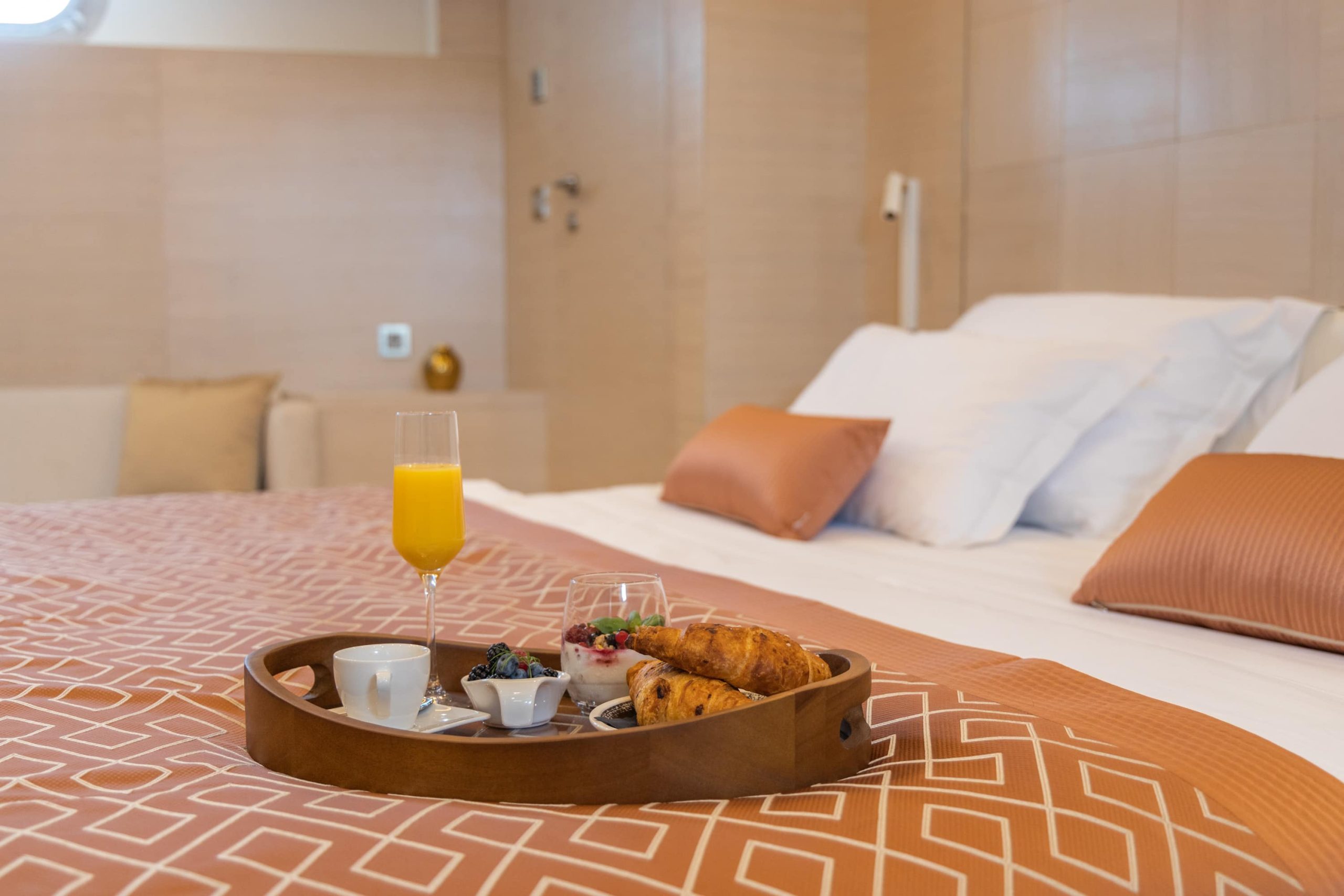 A wooden tray with breakfast, including croissants, fruit, yogurt, a cup of coffee, and a glass of orange juice, sits on a bed with patterned sheets and multiple pillows in a modern, cozy bedroom.