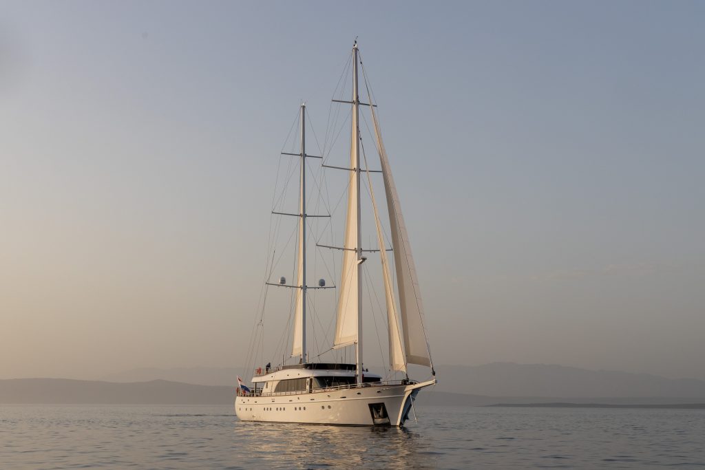 A large white sailboat with two tall masts sails on calm water at sunset, with distant mountains and a hazy sky in the background.