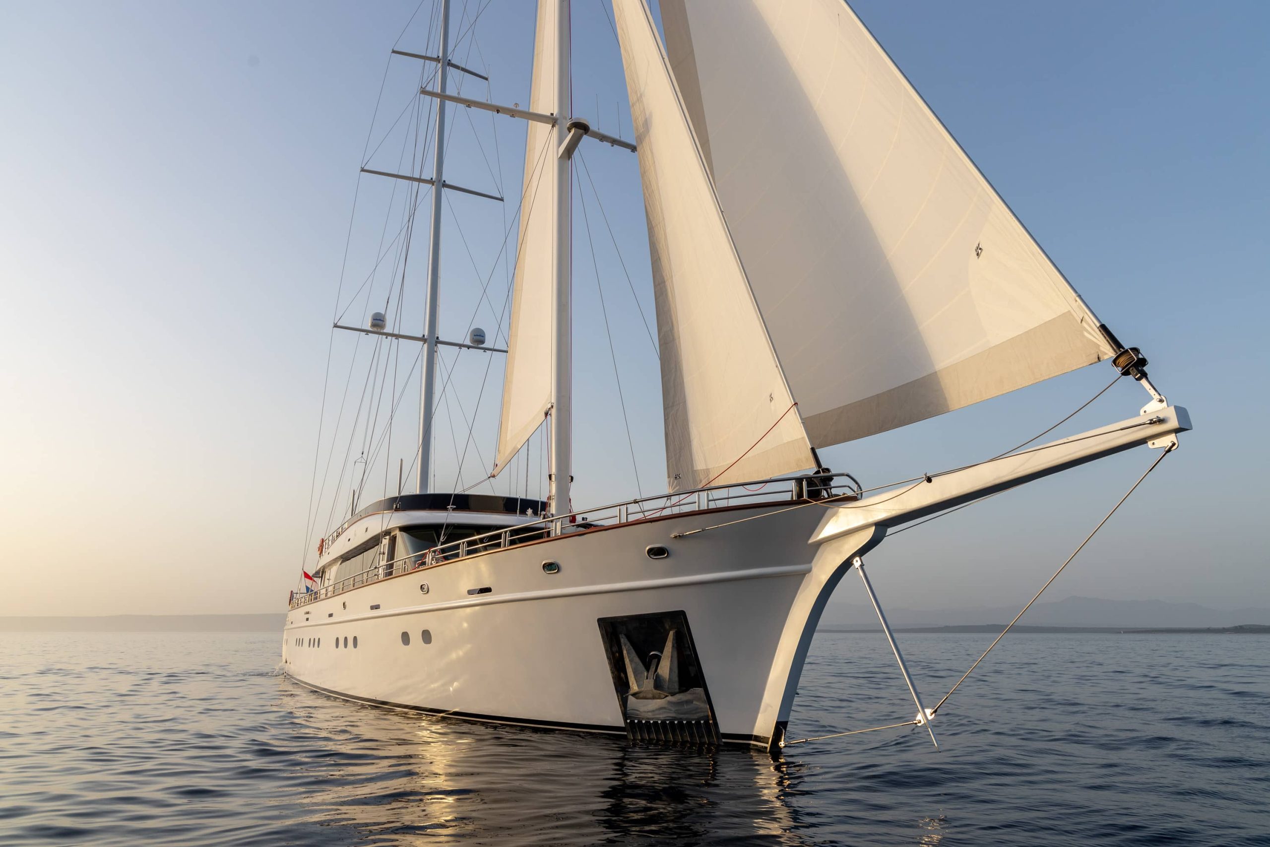 A large white sailing yacht with tall masts and full sails glides on calm water at sunset, with clear skies and distant land barely visible on the horizon.
