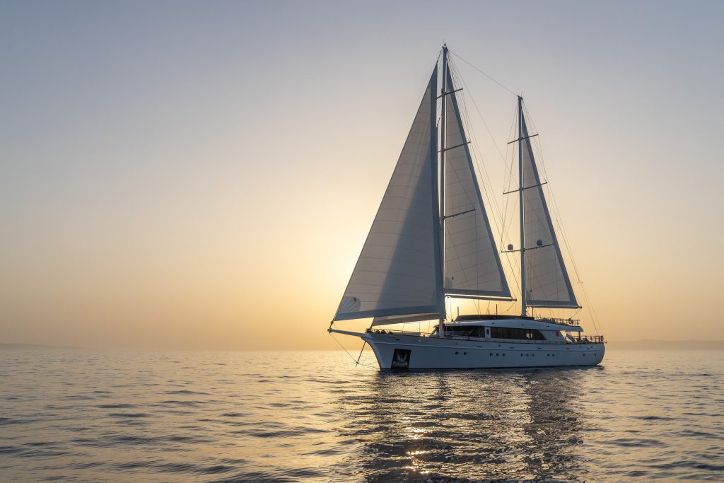 A large sailboat with two masts sails on calm water at sunset, with soft sunlight illuminating the sails and reflecting on the gentle waves.