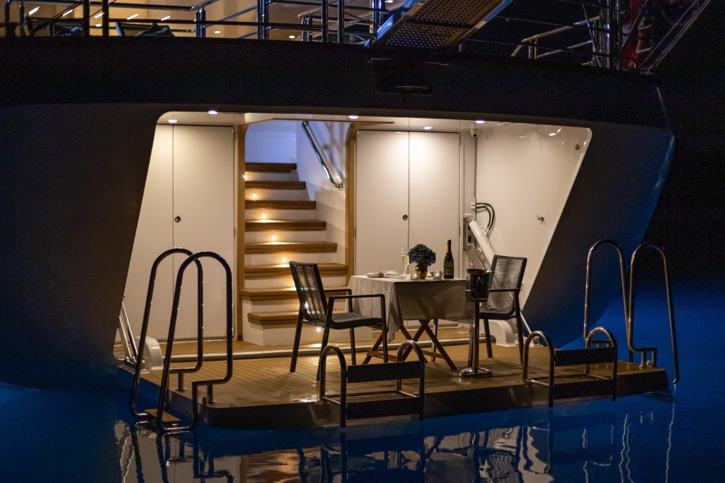 A luxury yacht’s illuminated aft deck at night, featuring a small dining table set for two with chairs, wine, and flowers beside a staircase, surrounded by water and metal railings.