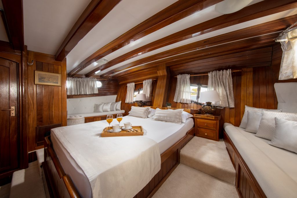 A cozy yacht cabin with wooden walls and ceiling beams, a large bed with white linens, a tray holding drinks, a cushioned bench, bedside tables, lamps, and windows with curtains letting in daylight.