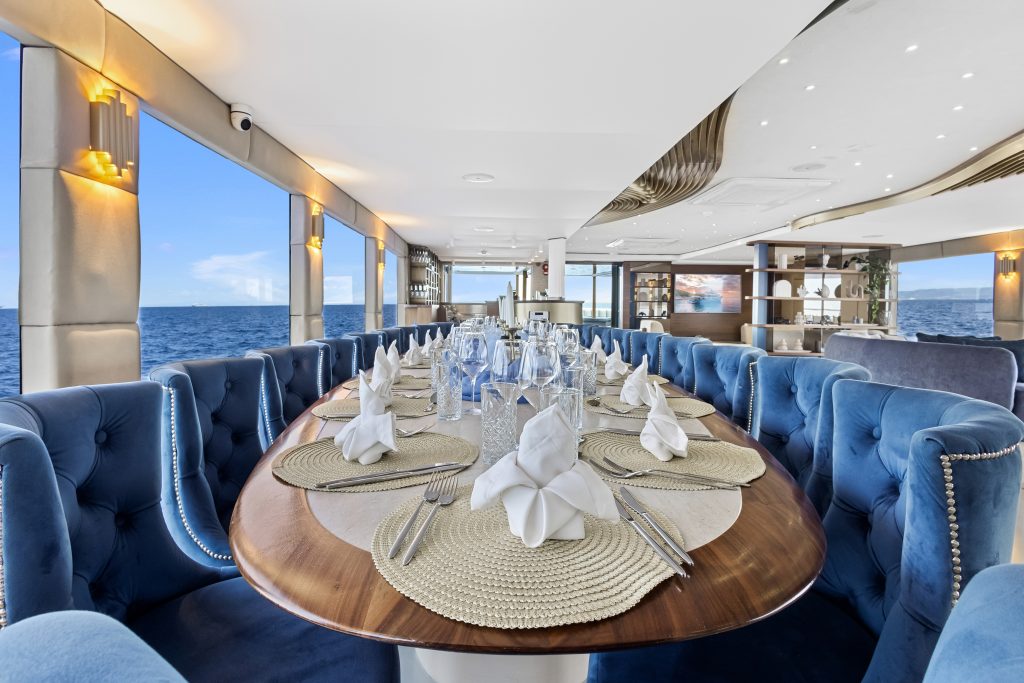 A luxurious dining area on a yacht with a long wooden table set for a meal, blue velvet chairs, and ocean views through large windows on both sides. The table features folded white napkins and glassware.