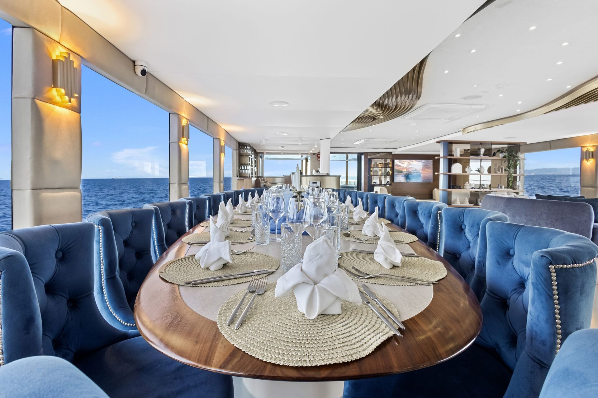 A luxurious dining area on a yacht with a long wooden table set for a meal, blue velvet chairs, and ocean views through large windows on both sides. The table features folded white napkins and glassware.