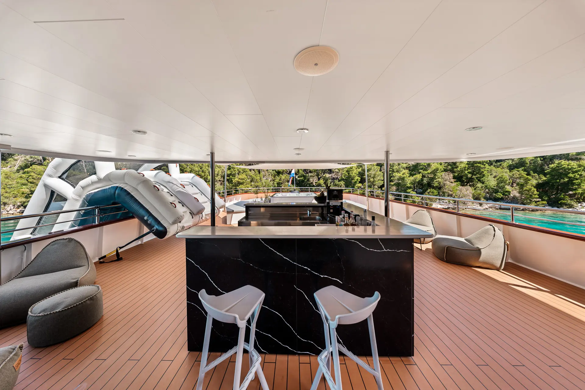 A modern yacht deck features a sleek black marble bar with two white chairs, lounge seating, and a view of green trees and blue water in the background. Inflatable slides extend over the railing.