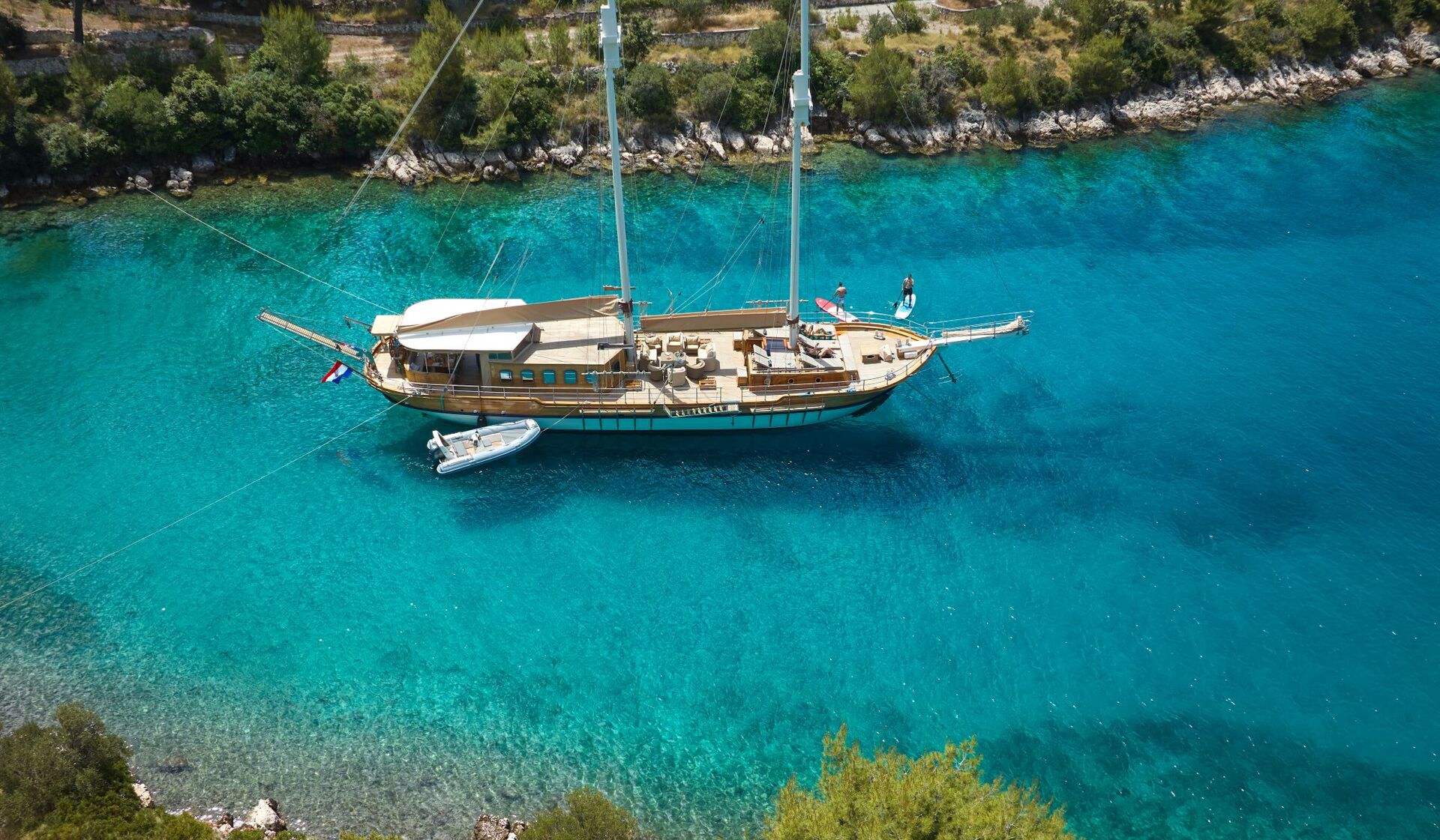LIBRA A wooden sailboat with two masts is anchored in clear turquoise water near a tree-lined rocky shore. A small inflatable dinghy floats beside the boat. The scene is viewed from above.