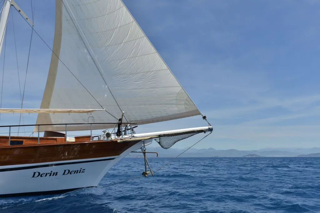 The bow of a wooden sailboat named Derin Deniz sails on calm blue water under a clear sky, with distant mountains visible on the horizon.
