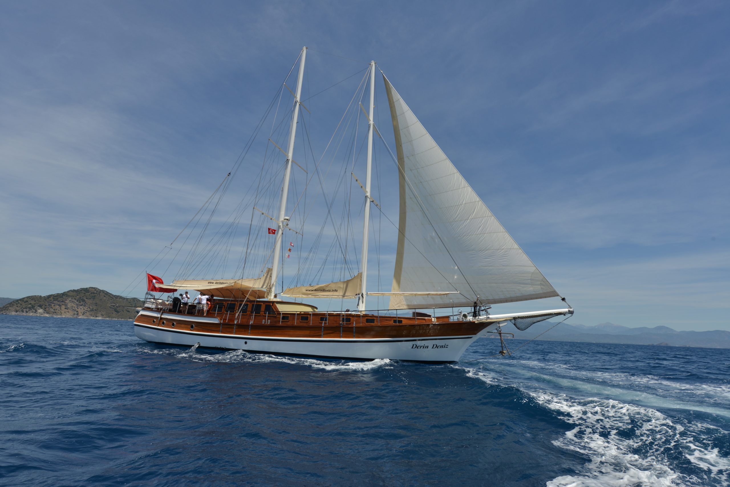 A large wooden sailing yacht with two masts glides on blue ocean water near a coastline, under a clear sky with scattered clouds. The Turkish flag is visible on the boat.