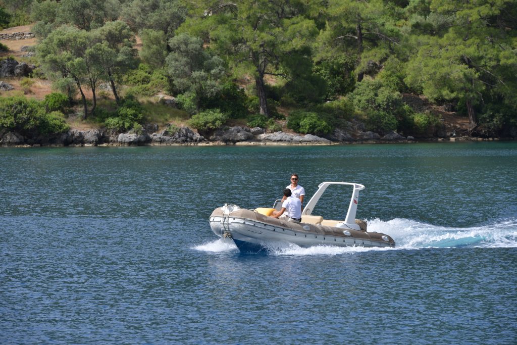Two people ride a small inflatable motorboat across calm blue water, with green trees and rocky shores in the background on a sunny day.