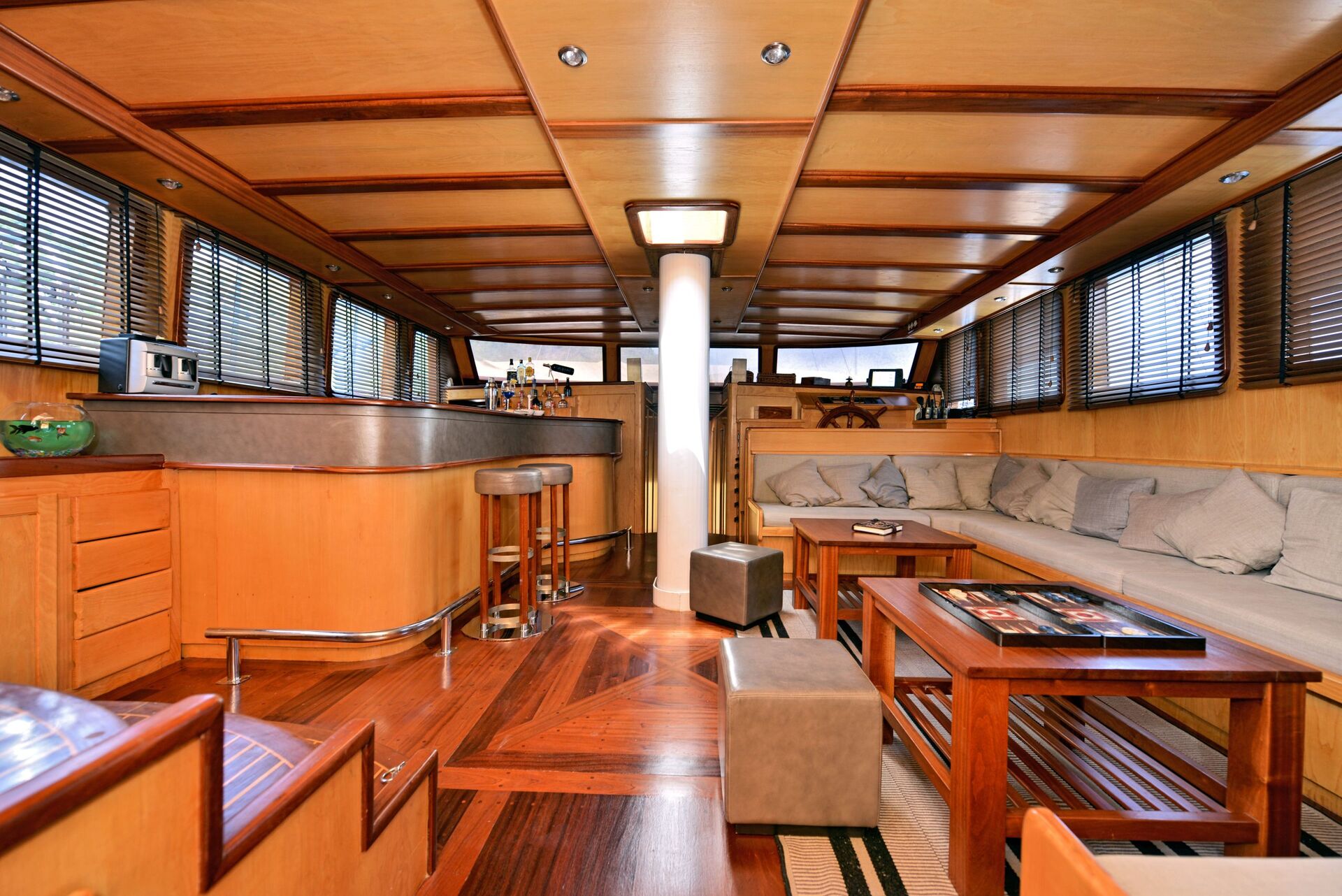Modern yacht interior with wood paneling, a long cushioned sofa, coffee table, ottomans, and a bar area with stools. Large windows with blinds let in natural light, giving the space an open, cozy feel.
