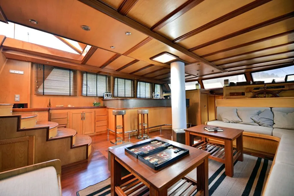 A spacious boat interior features wood paneling, a sitting area with a sofa and coffee tables, bar stools by a kitchen counter, and steps leading up to large windows letting in natural light.