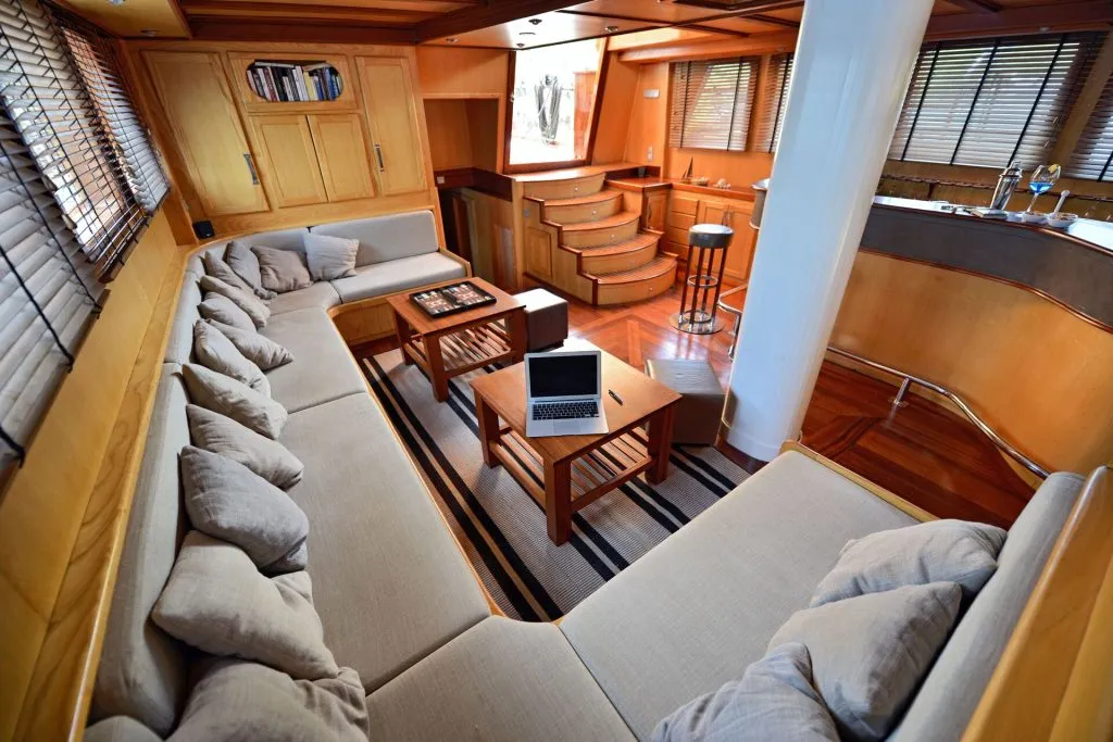 A spacious yacht interior featuring a U-shaped beige sofa with many pillows, wooden tables—one with a chessboard and laptop—polished wood floors, stairs, bookshelves, large windows, and a small kitchen area with a stool.