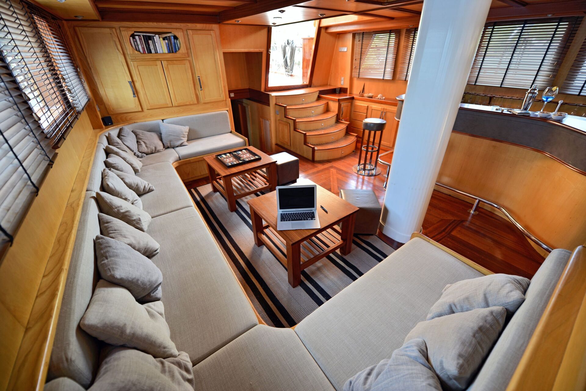 A spacious yacht interior featuring a U-shaped beige sofa with many pillows, wooden tables—one with a chessboard and laptop—polished wood floors, stairs, bookshelves, large windows, and a small kitchen area with a stool.