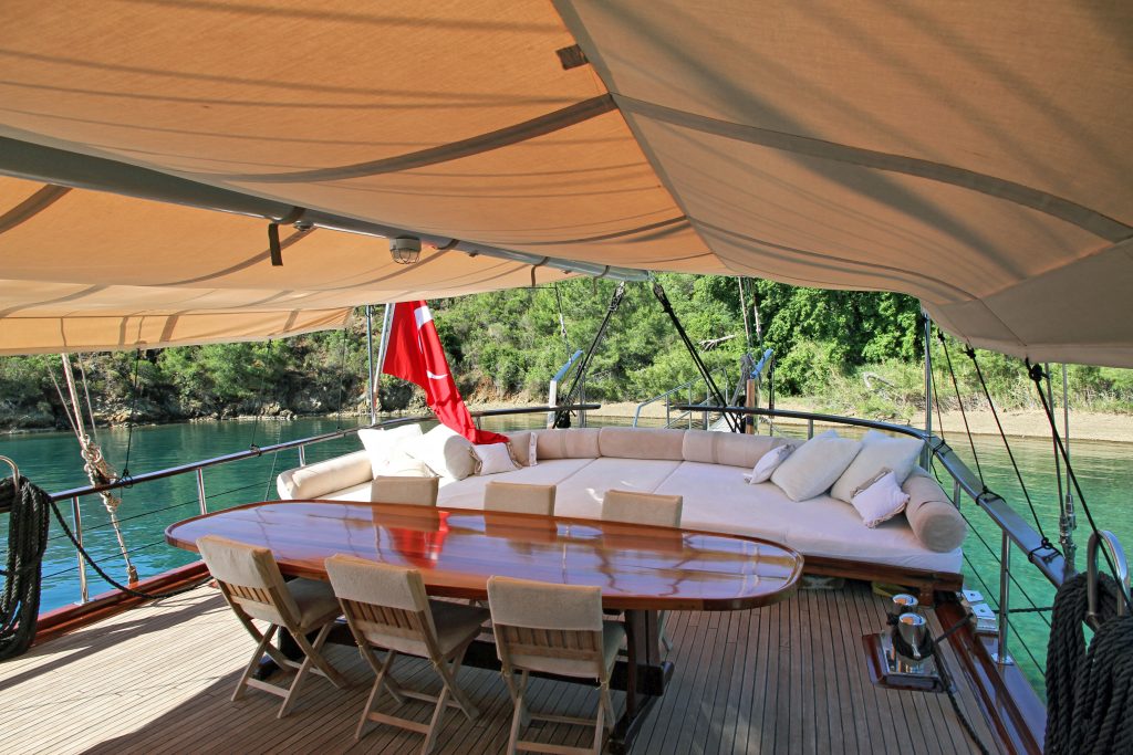 A covered outdoor yacht deck with a long wooden table, six chairs, a curved beige sofa with cushions, and views of calm water and lush green trees in the background. A red flag is visible at the back.