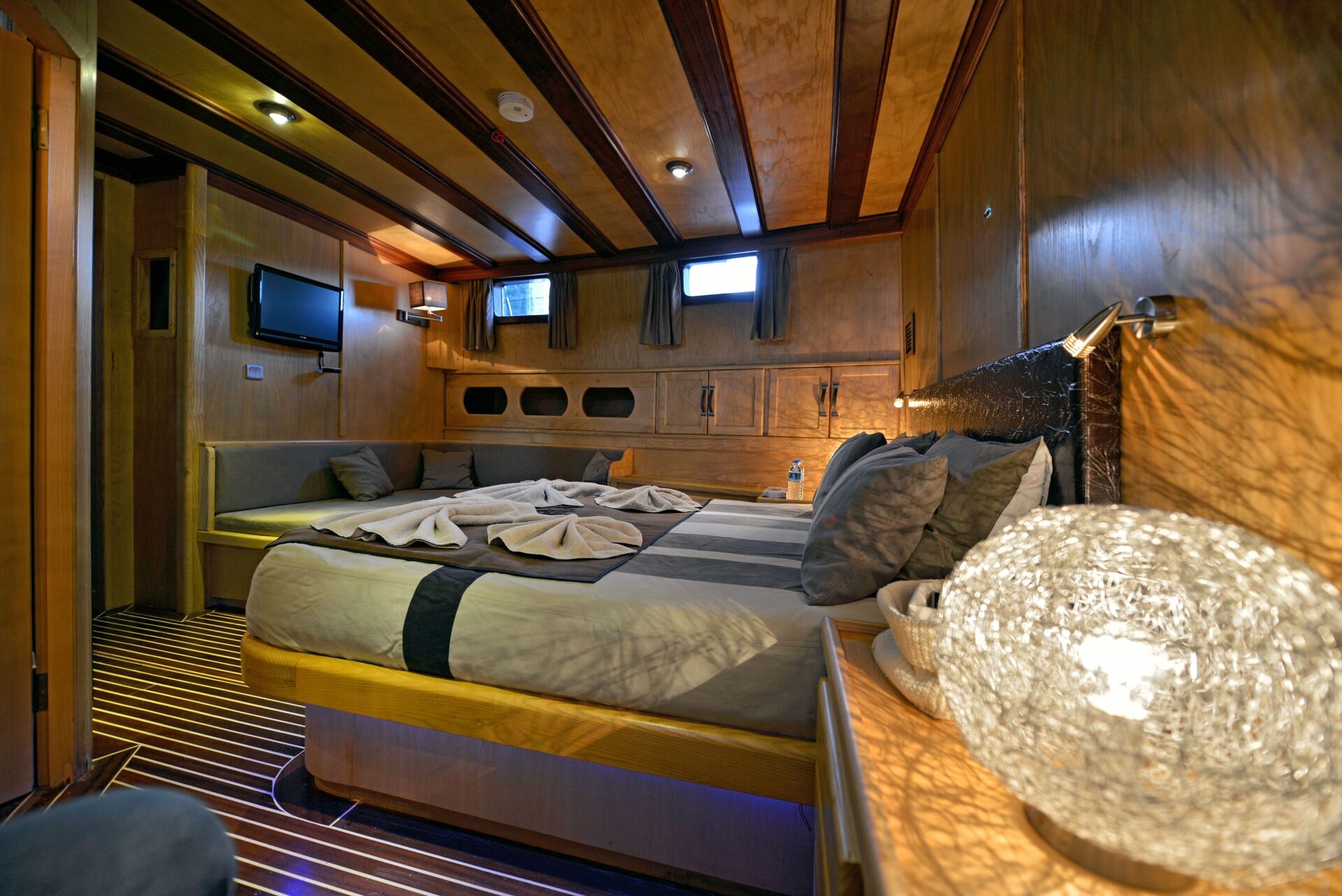 A cozy boat cabin with wood-paneled walls and ceiling, a queen-sized bed, sofa seating, small windows, a flat-screen TV, and a glowing decorative lamp on a side table. Soft lighting creates a warm ambiance.