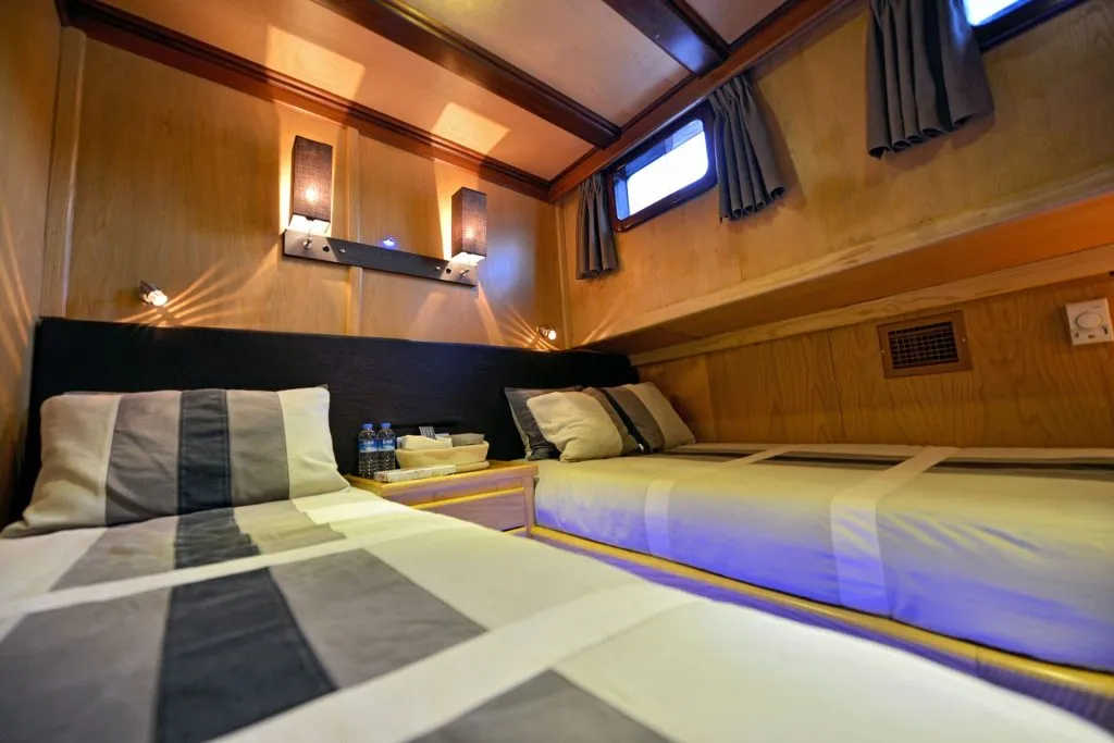 Cozy cabin with two single beds, striped bedding, a small nightstand with bottled water and tissues, wood-paneled walls, curtained windows, and soft overhead lighting creating a warm ambiance.