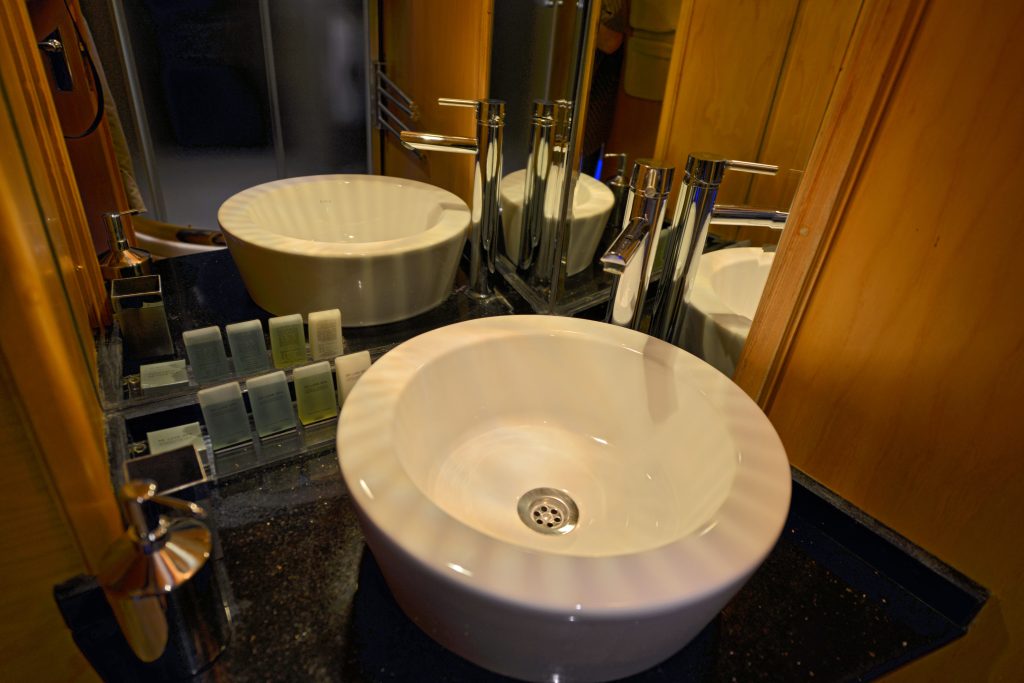 A modern bathroom sink with a round white basin, metal faucet, soap dispenser, and small toiletries arranged on a black countertop, reflected in a large wall mirror.