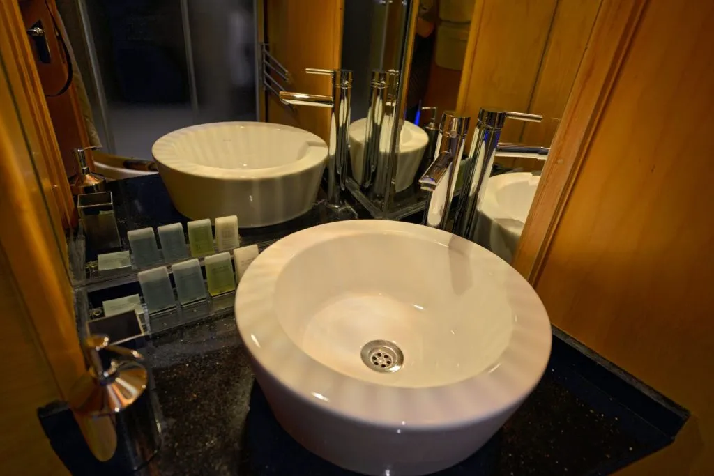 A modern bathroom sink with a round white basin, metal faucet, soap dispenser, and small toiletries arranged on a black countertop, reflected in a large wall mirror.
