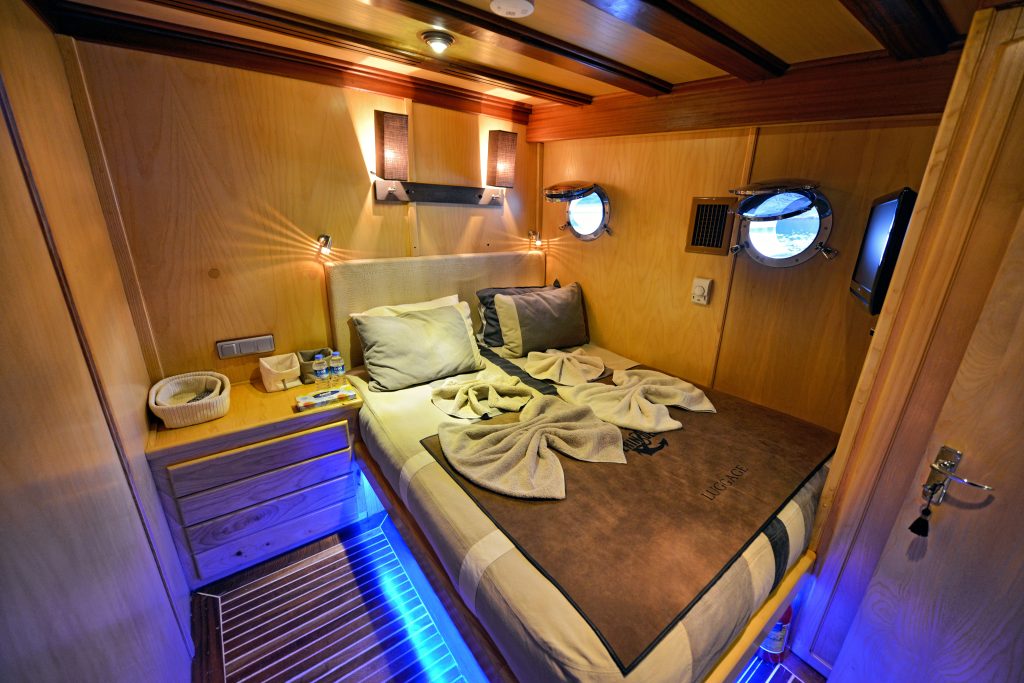 Cozy wooden cabin room with a double bed, neatly arranged towels, two porthole windows, soft lighting, a small TV, nightstands, and blue accent lights under the bed.