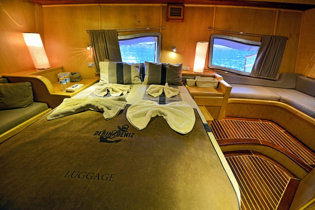 A neatly made bed with folded towel decorations in a cozy wooden cabin room, featuring large windows showing a water view, cushioned seating, soft lighting, and nautical-themed bed linens.