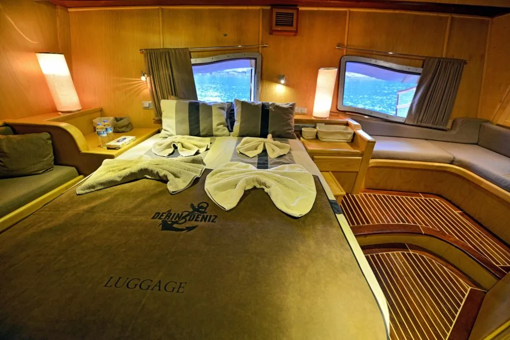 A neatly made bed with folded towel decorations in a cozy wooden cabin room, featuring large windows showing a water view, cushioned seating, soft lighting, and nautical-themed bed linens.
