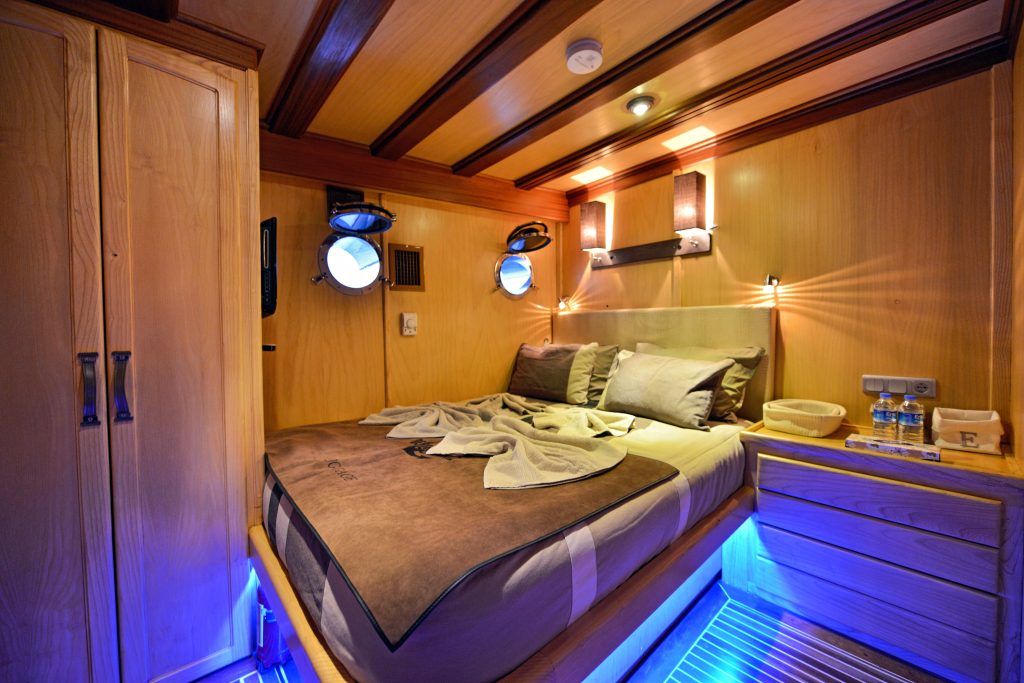 Cozy wooden cabin bedroom with a large bed, two round porthole windows, soft lighting, a towel folded on the bed, water bottles on the nightstand, and blue accent lights under the bed.