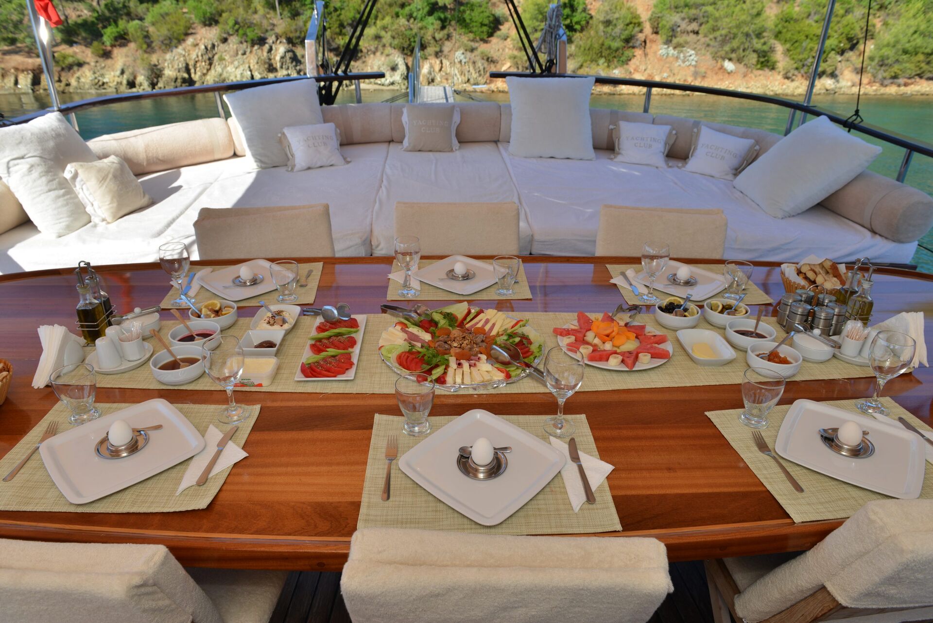 A table set for six with plates, cutlery, glasses, and egg cups, surrounded by platters of fruit, cheese, pastries, and appetizers on a yacht deck overlooking water and greenery.