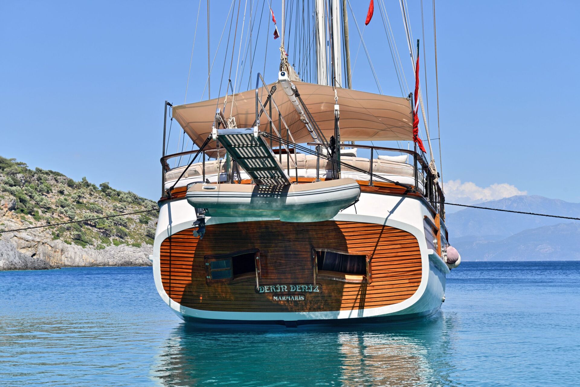 A wooden sailboat with tan canopies is anchored in calm blue water near a rocky coastline, with mountains and a clear sky in the background. The boat’s rear nameplate reads “Ceyru Deniz.”.