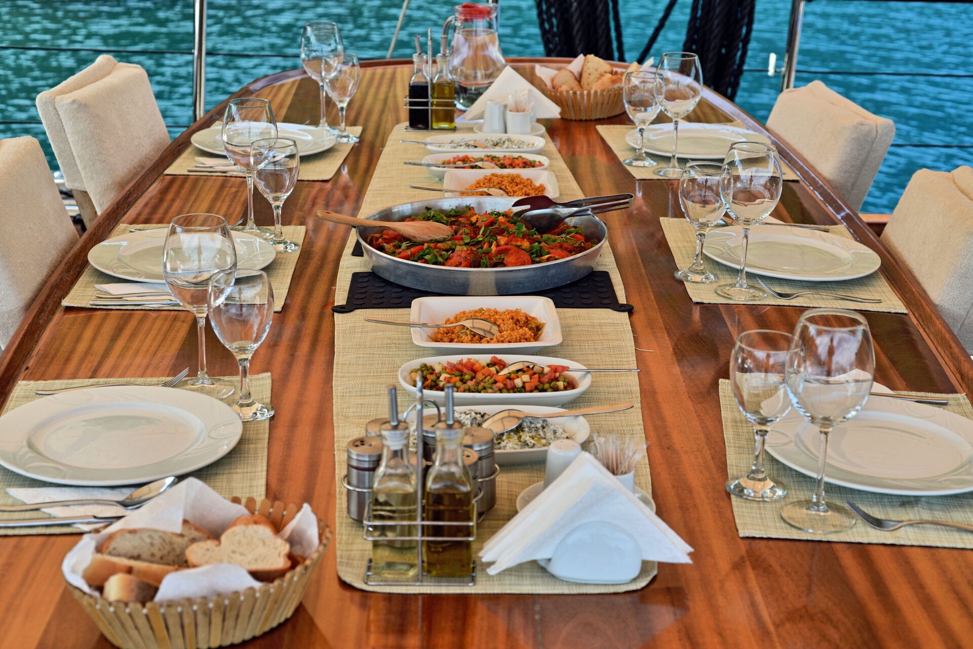 A wooden table set for a meal with plates, glasses, utensils, bread, olive oil, salads, and dishes of food, arranged neatly on placemats beside a body of water.