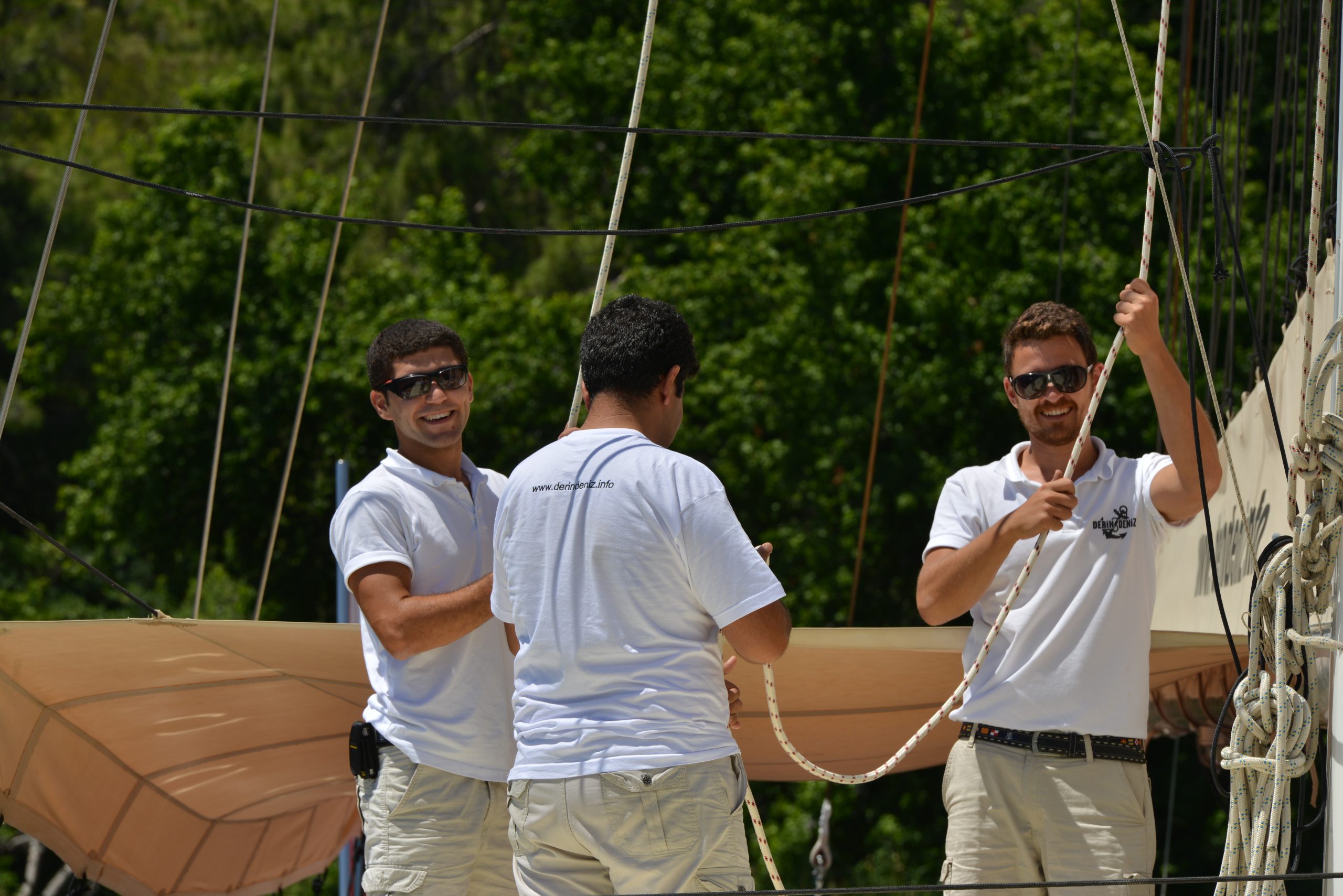 Three men in white polo shirts and sunglasses are handling ropes on a sailboat. Two are smiling at the camera, while one has his back turned. Green trees are visible in the background.