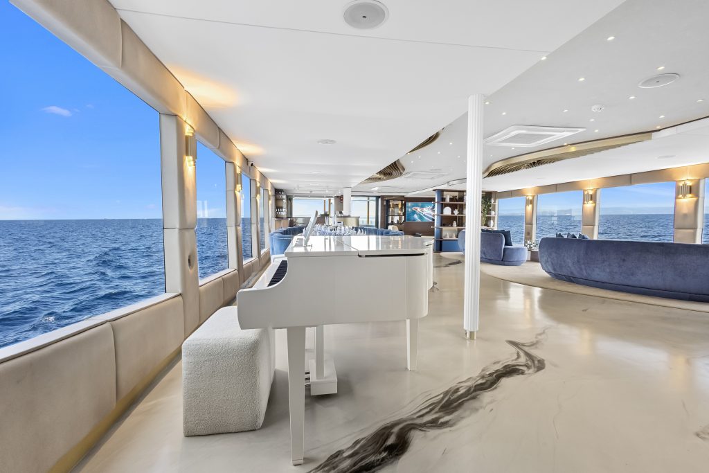 Luxurious yacht interior with large windows showing the ocean, a white grand piano, modern seating, and elegant marble-patterned floors, all bathed in natural light from the expansive sea views.