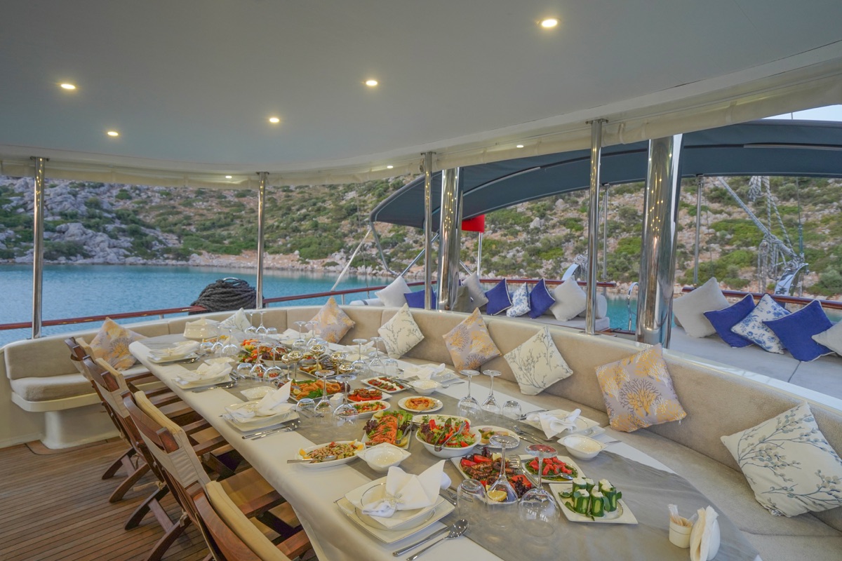 A luxurious outdoor dining area on a yacht features a long table set with various dishes and drinks, cushioned seating with decorative pillows, and scenic views of water and green hills in the background.