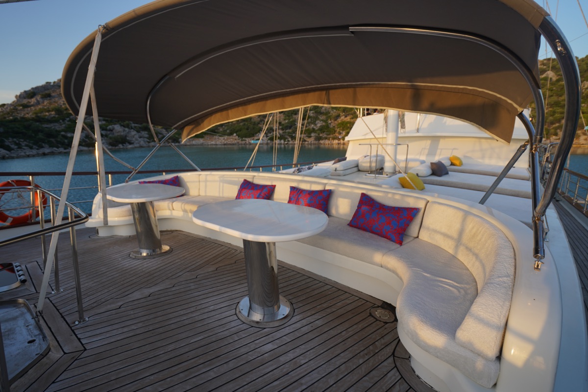A curved white cushioned seating area with colorful pillows and two round tables sits under a canopy on the deck of a yacht, with water and rocky hills in the background.