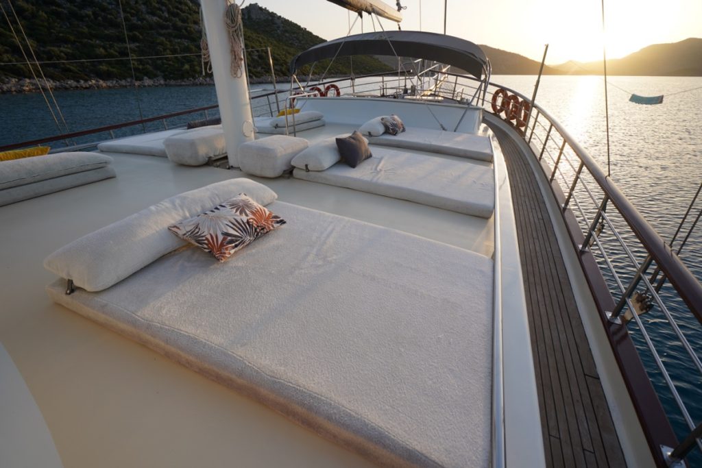 Sunlit deck of a yacht with cushioned lounge beds and decorative pillows, overlooking calm water and distant hills at sunset. A hammock hangs near the railing, creating a relaxing atmosphere.