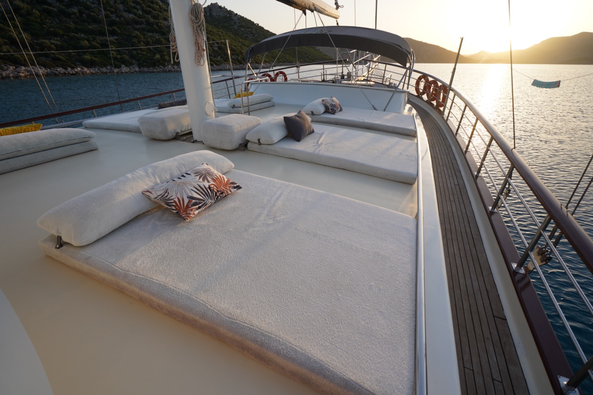 Sunlit deck of a yacht with cushioned lounge beds and decorative pillows, overlooking calm water and distant hills at sunset. A hammock hangs near the railing, creating a relaxing atmosphere.