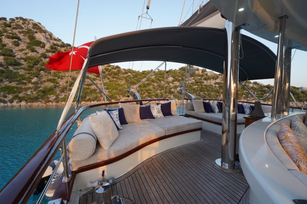 A luxury yacht deck with cushioned seating and decorative pillows, overlooking calm turquoise water and a hilly, green shoreline. A Turkish flag is visible at the stern, and the area is shaded by a canopy.