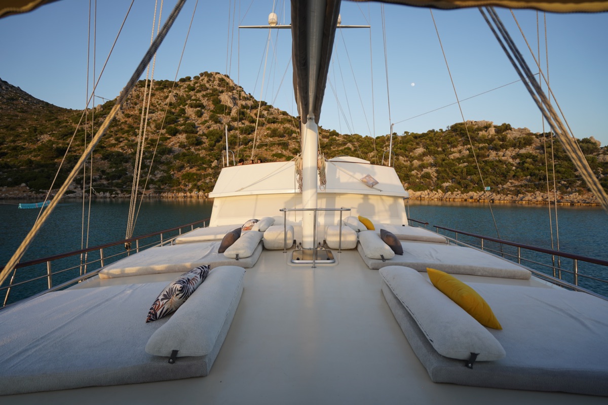 The deck of a yacht with cushioned lounge beds and colorful pillows, anchored near rocky, green hills under a clear sky at sunset.