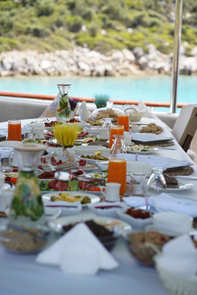 A table set for a meal on a boat, featuring various dishes, glasses of orange juice, and decorative items, with a scenic view of water and rocky shoreline in the background.