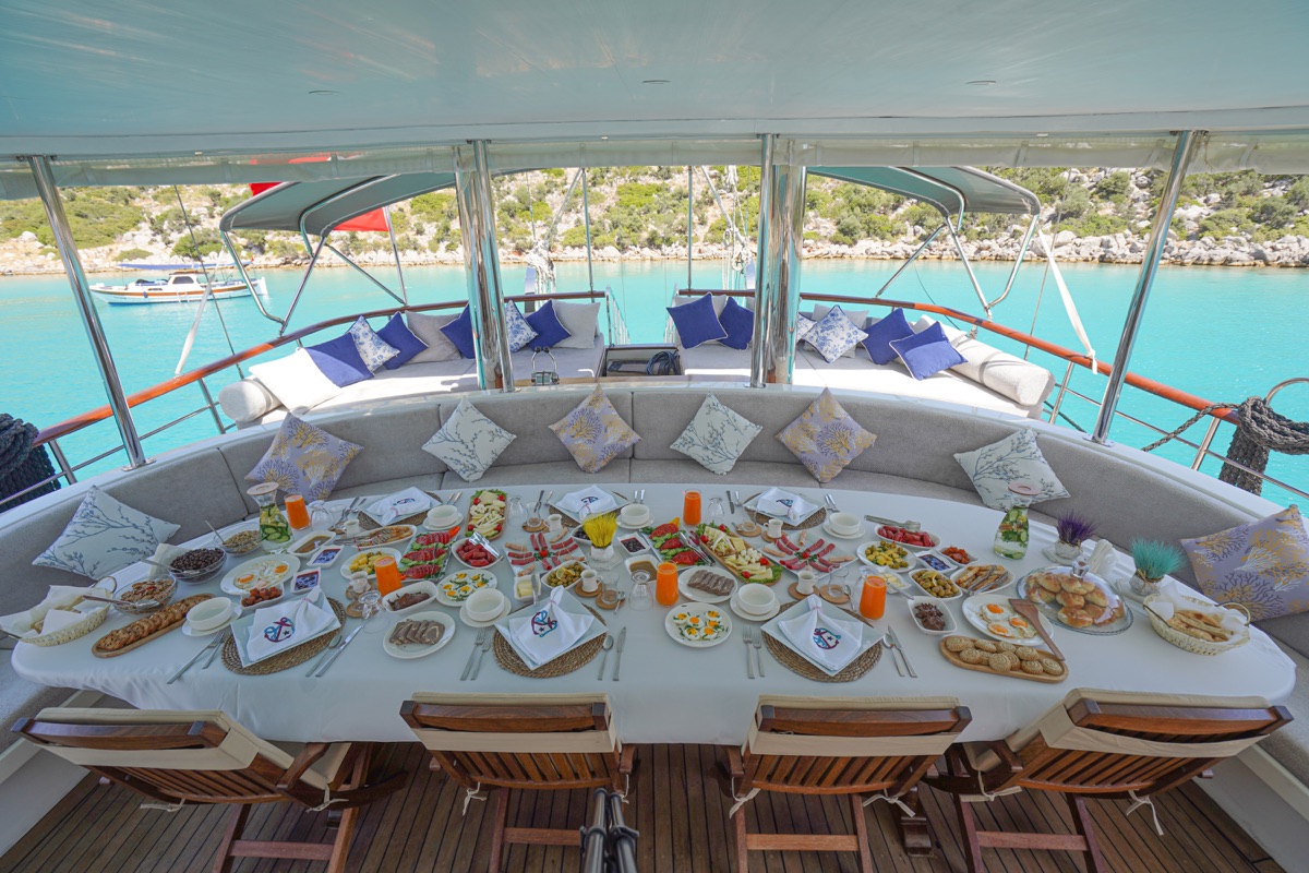 A luxurious yacht deck set for a meal with an elegant white tablecloth, plates of food, drinks, and wooden chairs. Cushioned seating and decorative pillows line the back, with turquoise water and boats visible in the background.