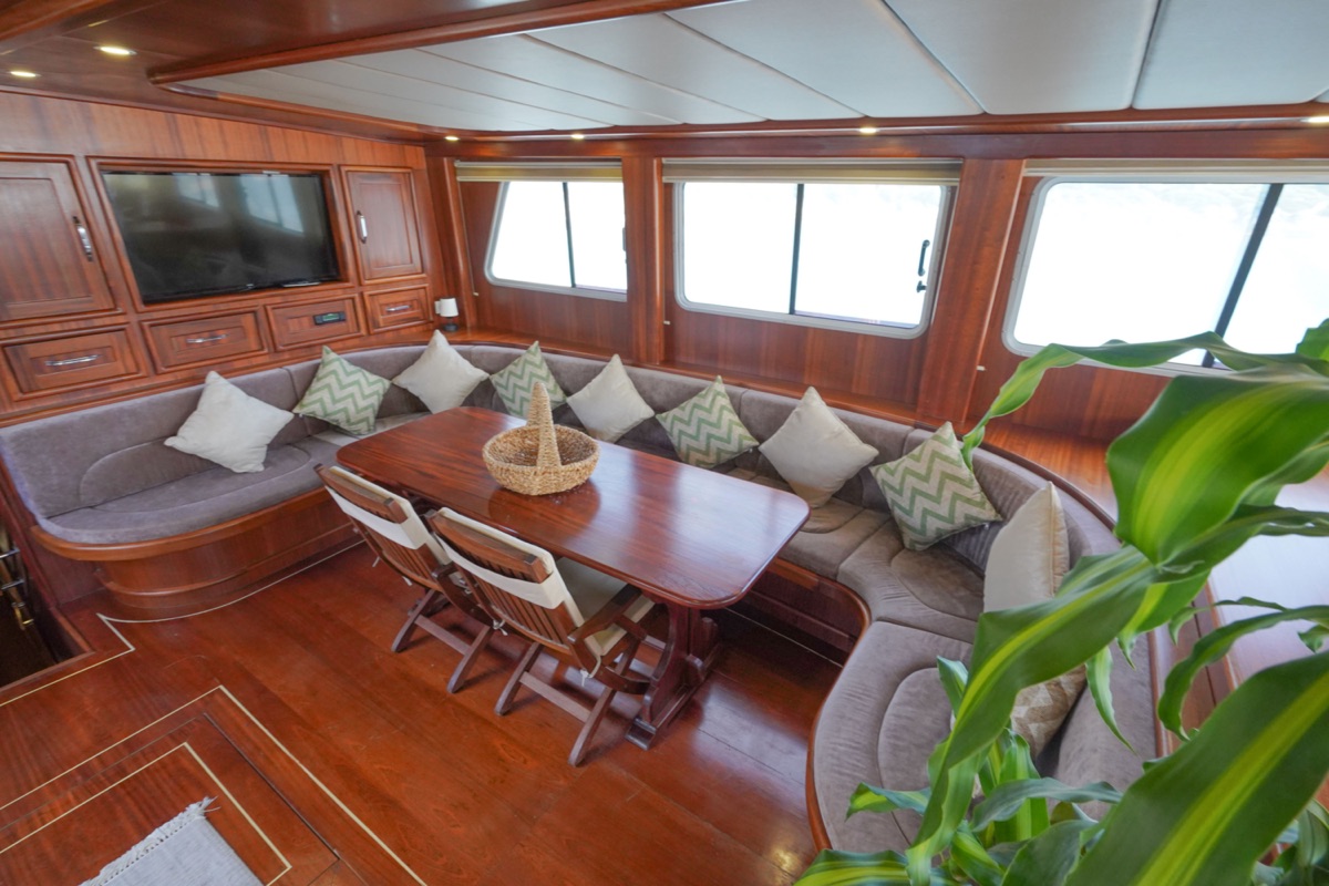 A cozy yacht lounge with a curved sofa, decorative pillows, a polished wooden table, two chairs, a wall-mounted TV, large windows, and a green plant in the corner. The space features rich wood paneling throughout.