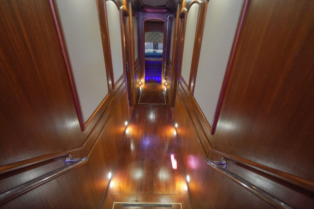 A polished wooden hallway with handrails and recessed lights along the floor, leading to a room with a visible bed and blue lighting at the far end.
