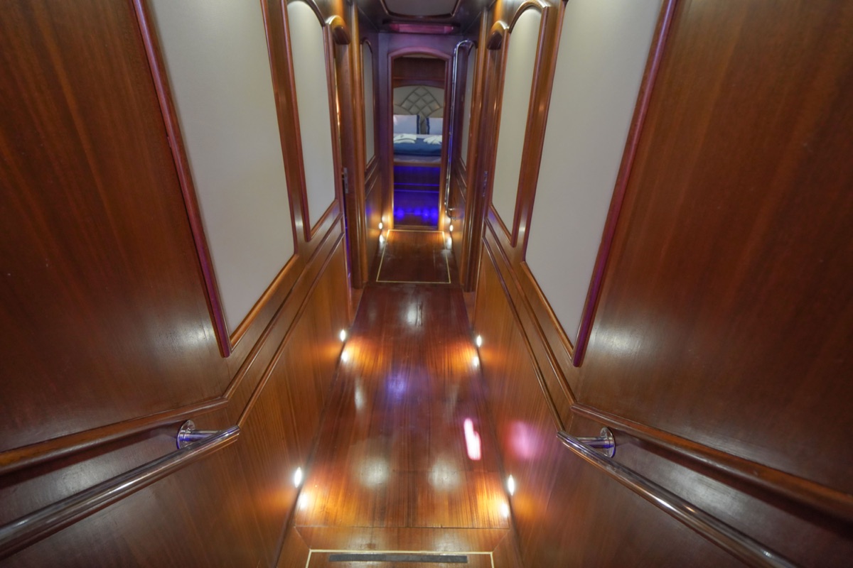 A polished wooden hallway with handrails and recessed lights along the floor, leading to a room with a visible bed and blue lighting at the far end.