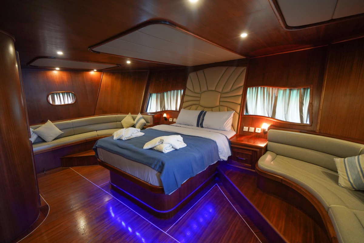 Luxurious yacht cabin with wood-paneled walls, a central bed with blue and white linens, two curved sofas, soft lighting, and blue accent lights illuminating the polished wooden floor.