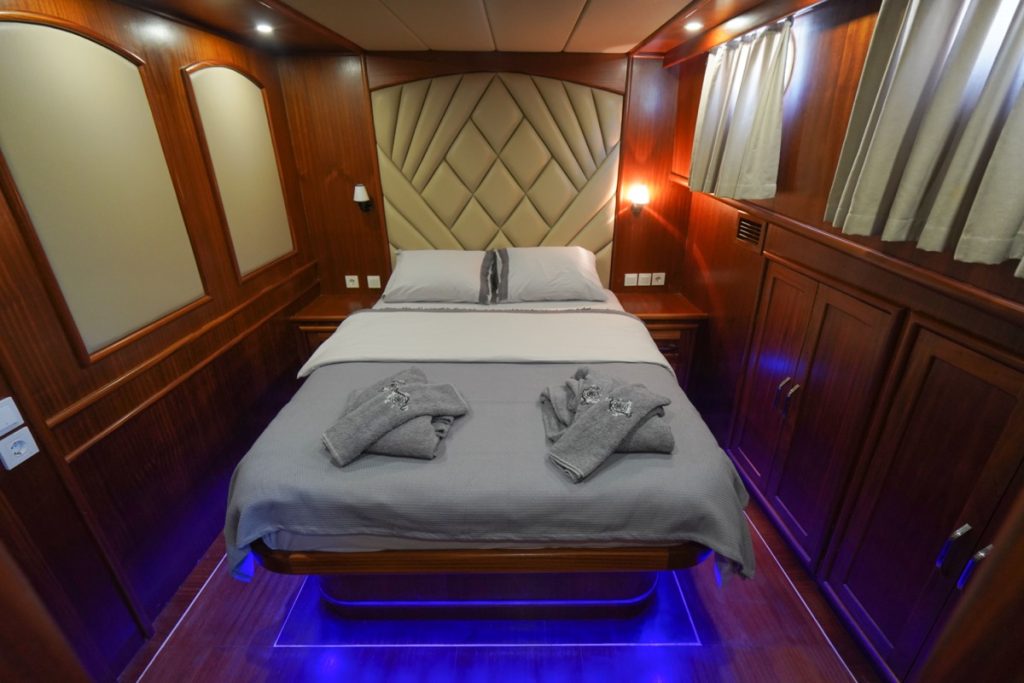 A neatly made bed with grey linens and folded towels is centered in a compact, wood-paneled room with overhead lights, beige padded headboard, and curtains on the windows. Blue lights illuminate the floor below the bed.