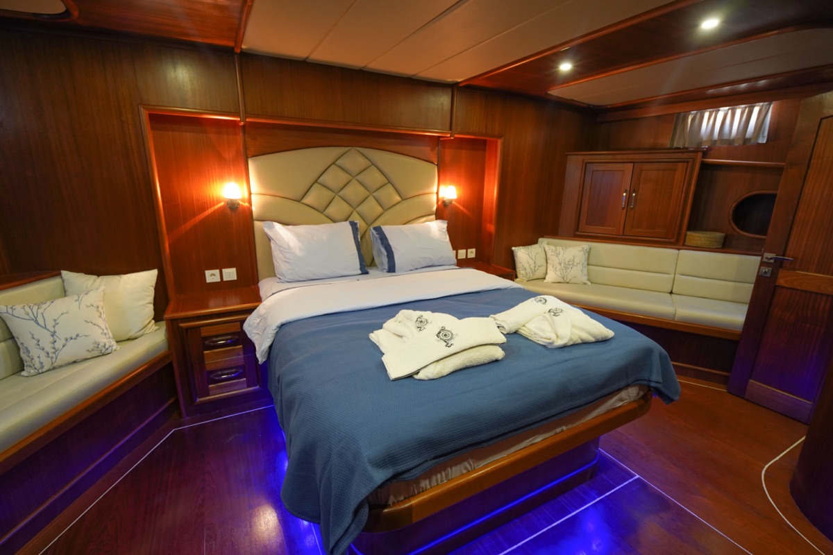 Luxurious bedroom with a large bed, blue blanket, white pillows, and folded towels. The room features wood-paneled walls, ambient lighting, a built-in sofa, and a modern, cozy atmosphere.