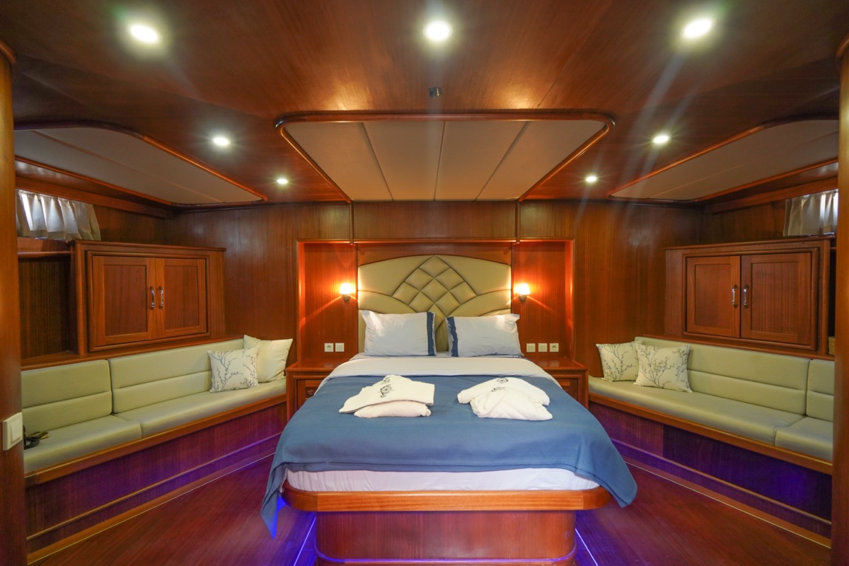 A spacious bedroom with rich wooden walls and ceiling, a large bed with blue bedding, two bathrobes laid out, cushioned benches along the walls, and warm ambient lighting.